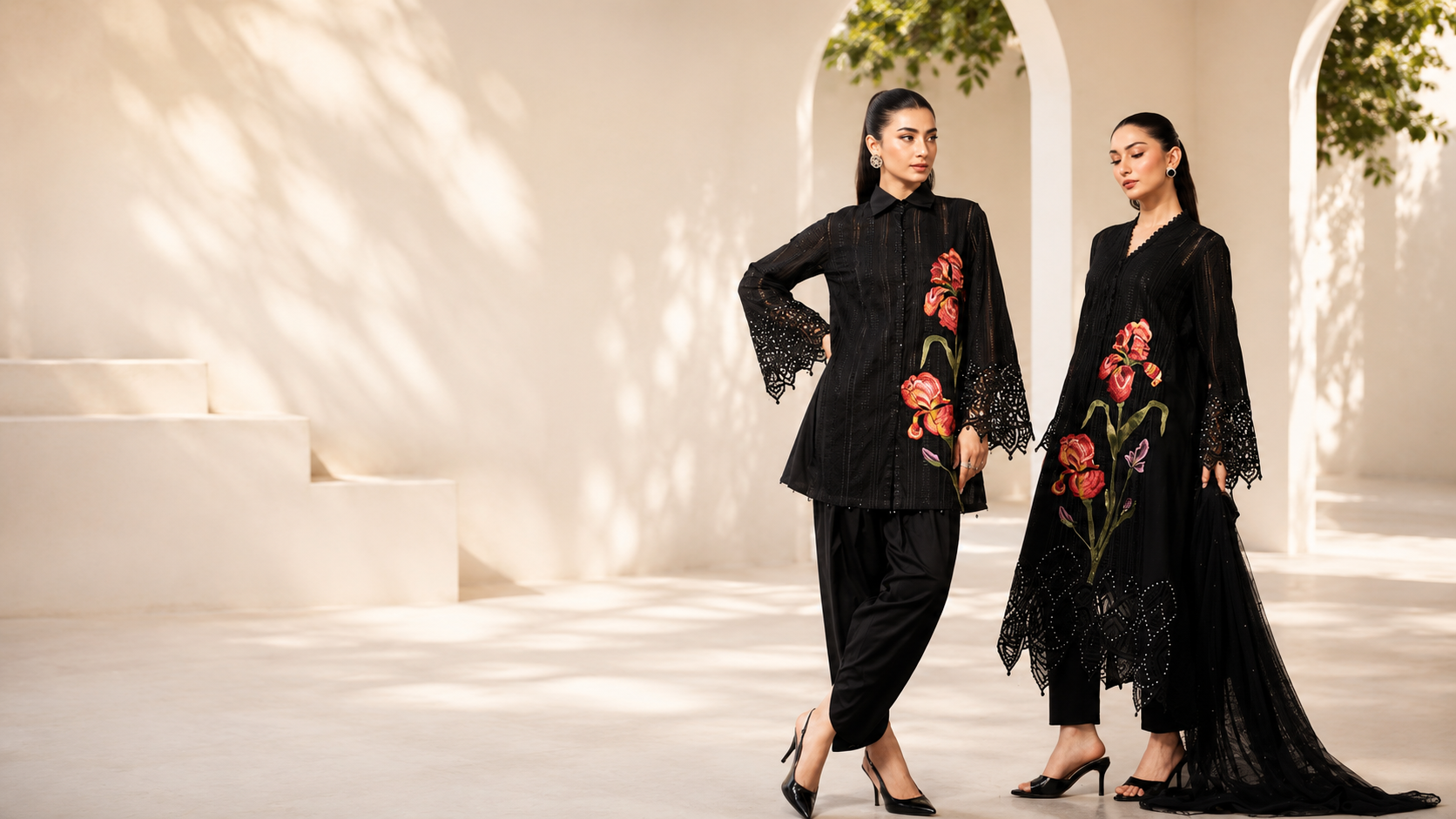 MK Signature — two models in black Pakistani designer suits with floral embroidery in a sunlit colonnade